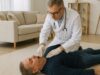 Ein Arzt in weißem Kittel und mit Stethoskop untersucht eine Person, die auf einem Teppich in einem Wohnzimmer liegt. Im Hintergrund sind ein Sofa, Kissen und ein Regal zu sehen.