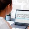 Eine Person sitzt vor einem Laptop, auf dessen Bildschirm das Programm „Hautkrebs-Screening Fortbildungsprogramm“ angezeigt wird. Im Hintergrund ist eine helle Wand und eine kleine Pflanze zu sehen. Die Person hält einen Stift in der Hand.