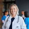 Blonde Ärztin mit Stethoskop und Nasenring schaut lachend in die Kamera. Im Hintergrund ist weiteres medizinisches Personal zu sehen.